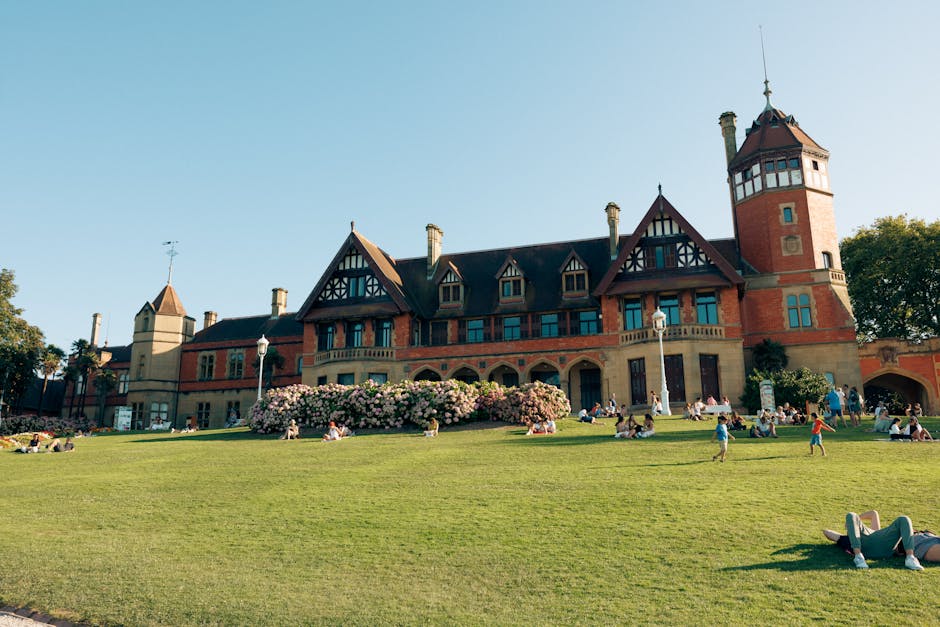 The image features a historic red brick building with multiple gabled roofs, turrets, and tall chimneys, set against a clear blue sky. In the foreground, there is a well-maintained, lush green lawn wi